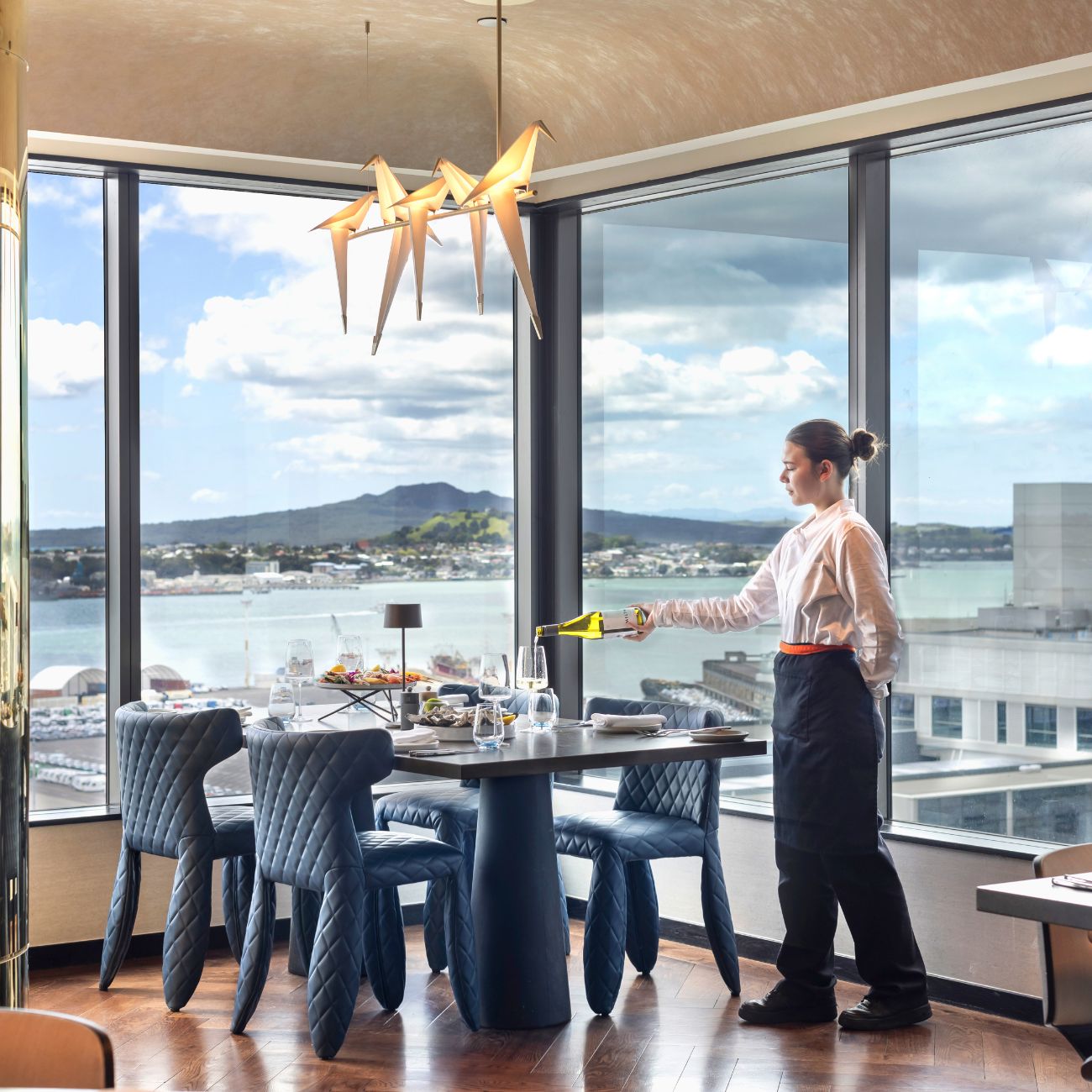 A waiter serves wine at a stylish restaurant table with panoramic views of a city skyline and water, decorated with modern furniture.