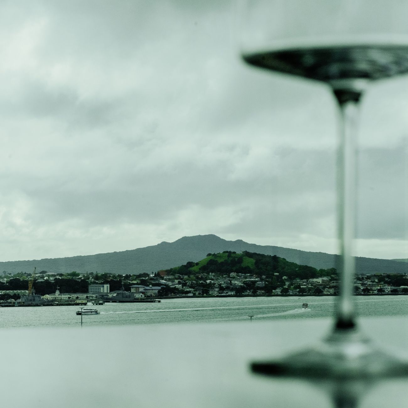 A glass of wine in the foreground frames a scenic view of a cloudy harbor and a distant hill across the water.