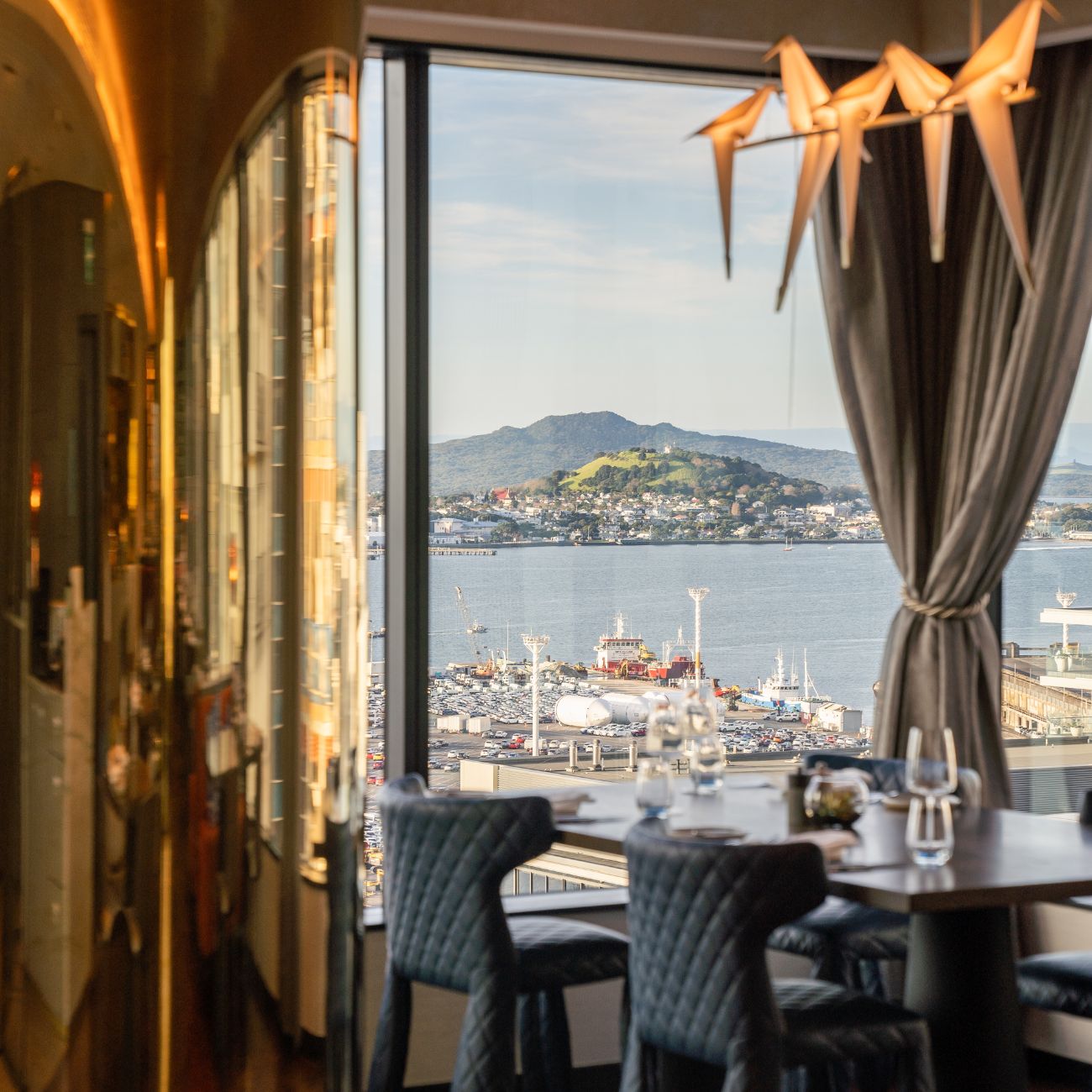 A stylish restaurant interior with a view of a harbor and lush hills in the background, featuring elegant seating and decorative lighting.