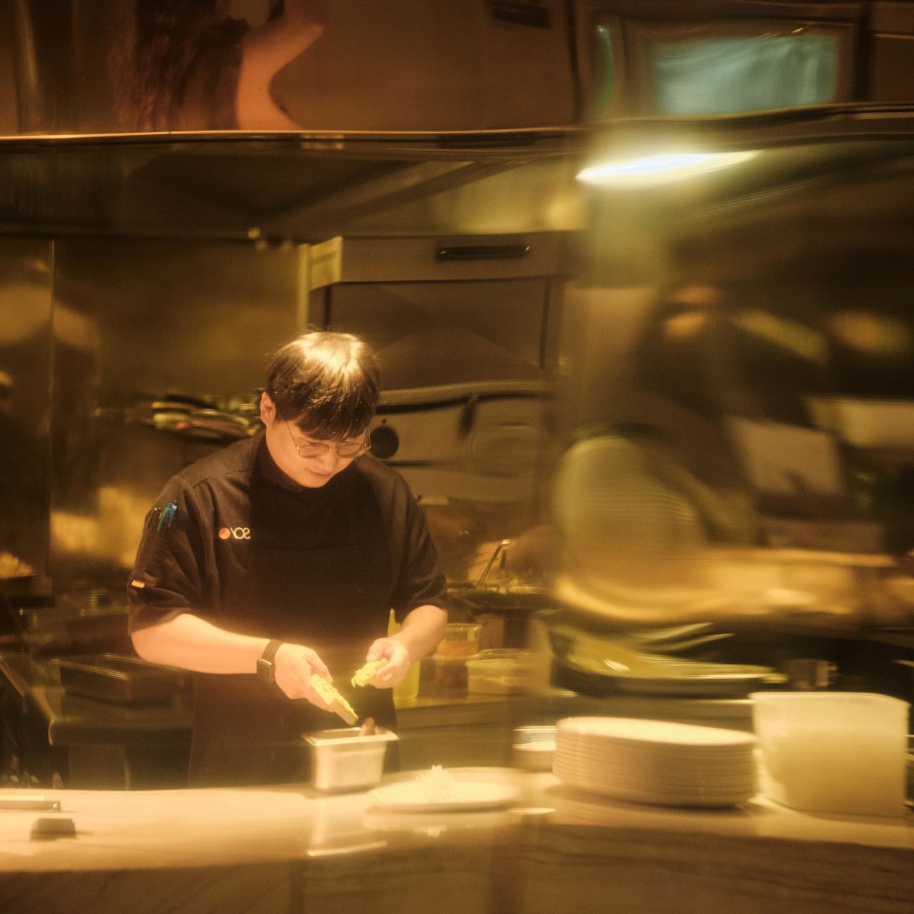 A chef prepares food in a shiny, modern kitchen, reflecting warm light from polished surfaces and organized kitchenware.