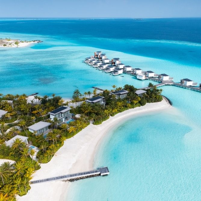 Aerial view of a tropical resort with overwater villas surrounded by turquoise waters and white sandy beaches lined with palm trees.