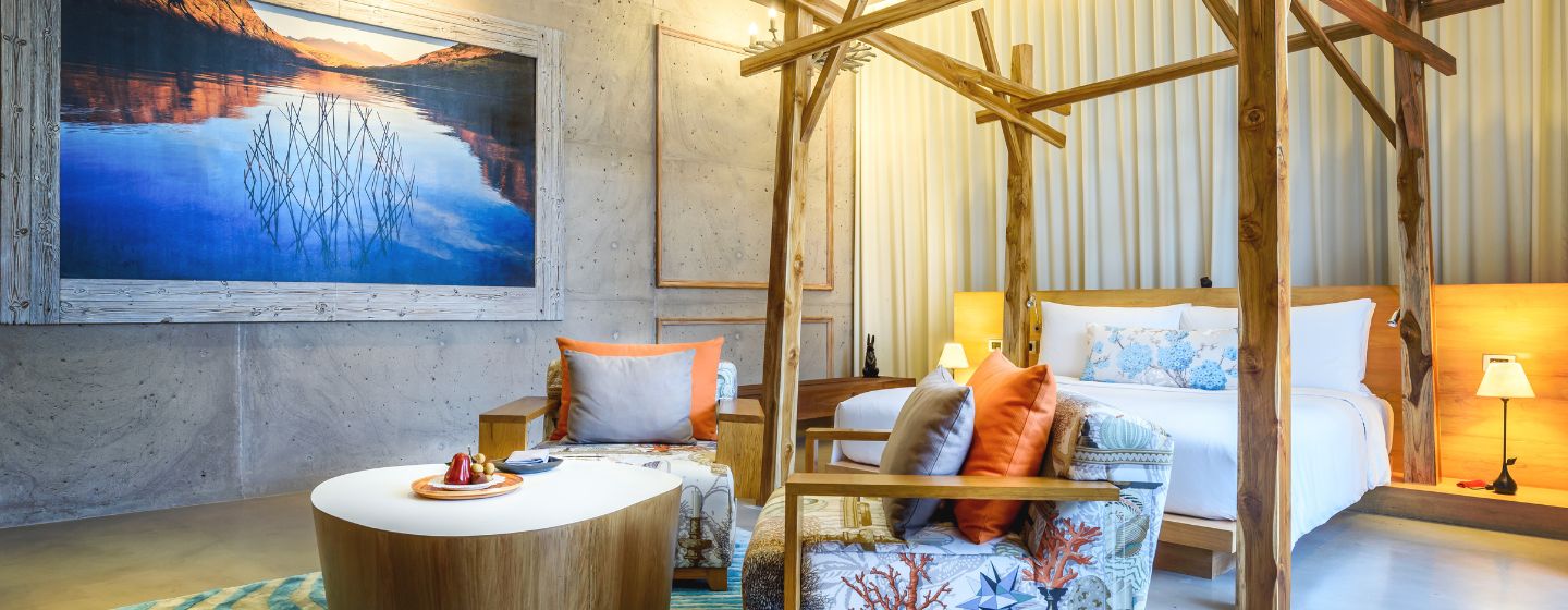 A cozy hotel room features a king-sized bed, vibrant cushions on a sofa, warm lighting, and nature-inspired decor, creating a serene ambiance.