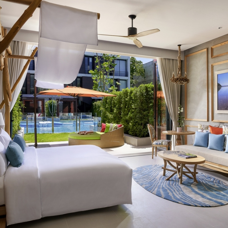 Modern, airy hotel room with a white bed adorned with teal pillows. Bamboo decor frames the headboard. A glass door leads to a balcony with chairs and a city view. The room features a ceiling fan and a light wood coffee table with fruit, evoking a calm, inviting atmosphere.