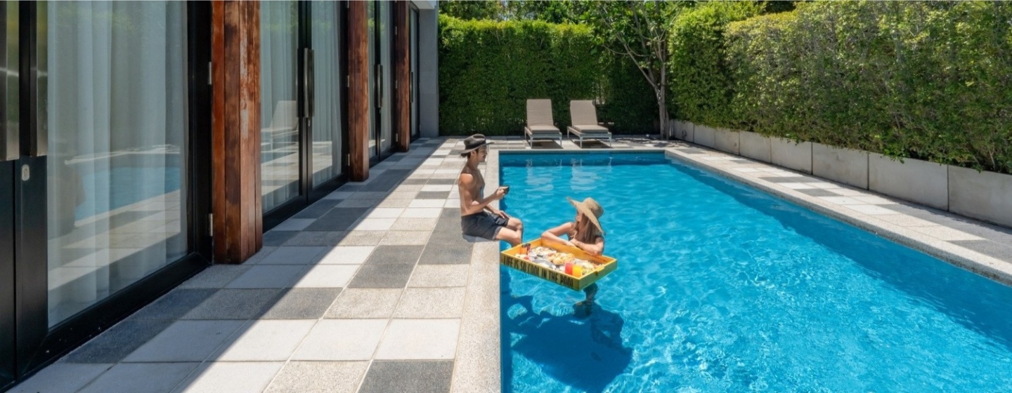 A serene backyard pool scene with a person sitting on the edge, another floating on a small raft. Sunlight filters through lush greenery surrounding the area.