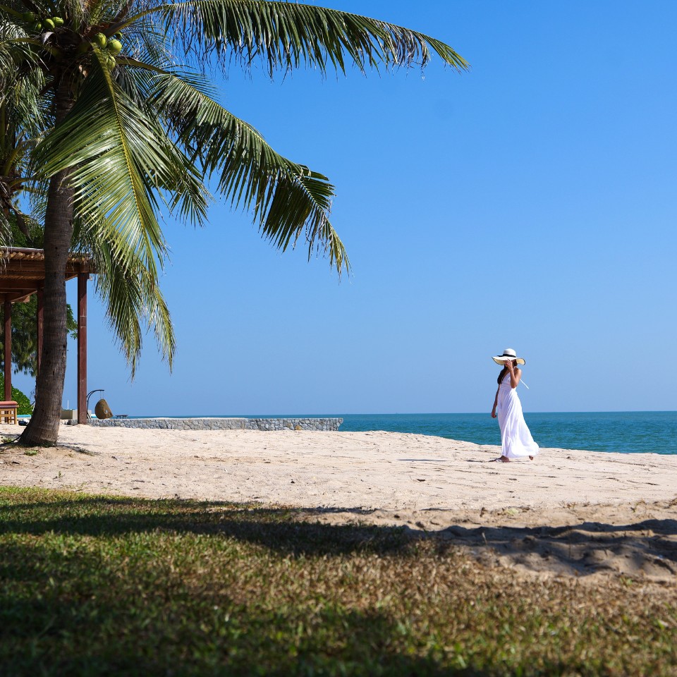 A woman in a white dress and hat strolls on a tropical beach by the ocean. Palm trees and a shaded lounge area create a serene, relaxed atmosphere.