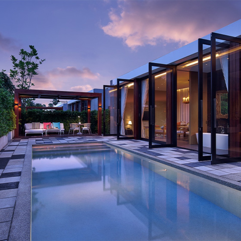 Modern house exterior at dusk with open glass doors, a lit interior, pool in the foreground, and greenery surrounding the patio area, creating a serene atmosphere.