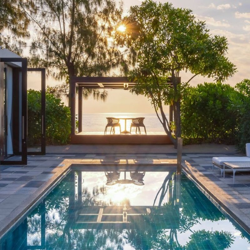 Sunset over a tranquil pool and ocean, framed by lush trees and a modern patio with chairs. Reflective water adds a peaceful, relaxing vibe.
