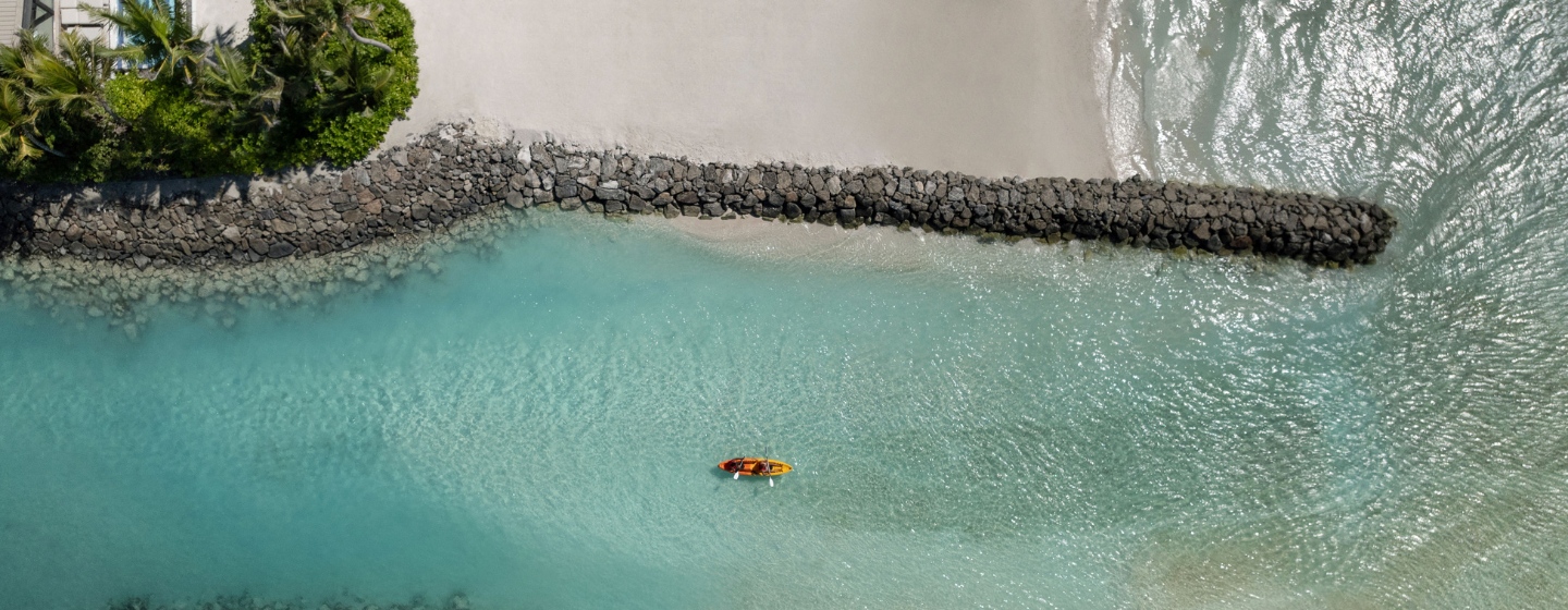 Aerial view of a kayak on turquoise water near a sandy beach and rock jetty, surrounded by lush greenery.