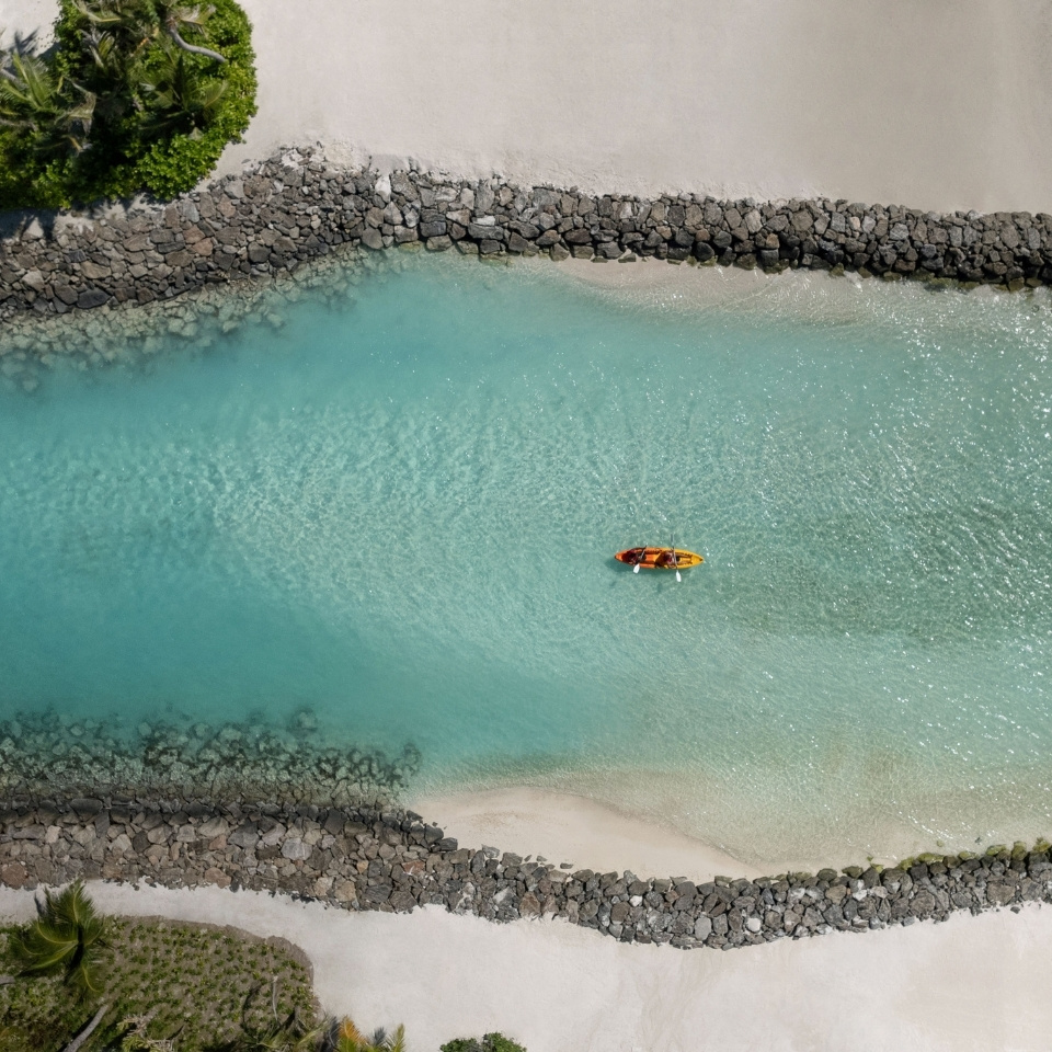 Aerial view of a kayak on turquoise water near a sandy beach and rock jetty, surrounded by lush greenery.