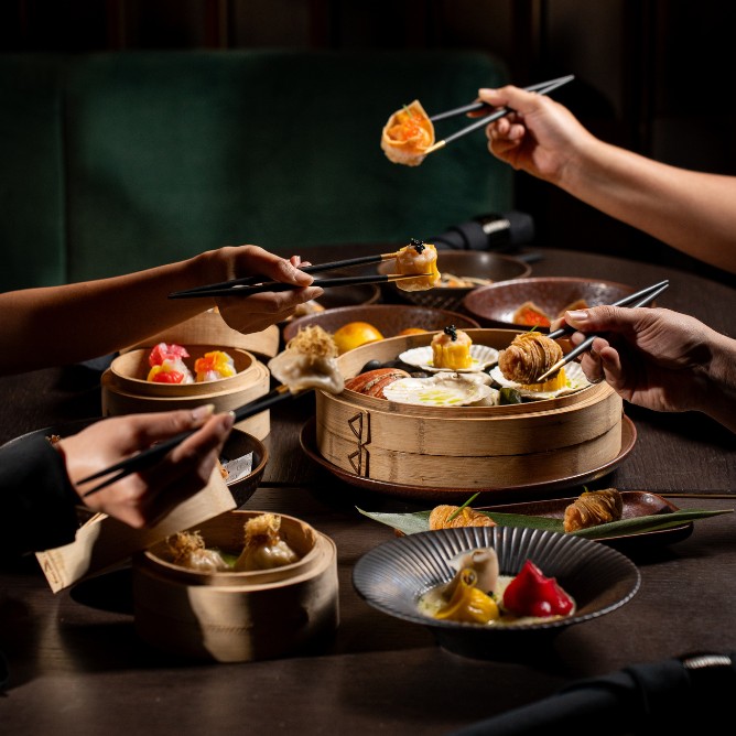 A variety of dumplings and dim sum are being enjoyed with chopsticks by multiple hands at a stylish dining table.