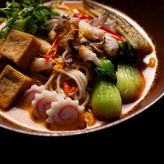 A vibrant bowl of noodle soup with seafood, bok choy, tofu, and colorful toppings, garnished with fresh cilantro and sliced chilies.