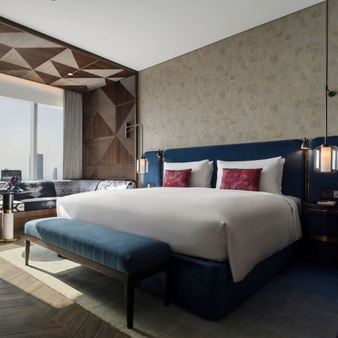 A modern hotel room with a king-sized bed, white linens, and red pillows. The room features geometric wood paneling, large window, and a city view.