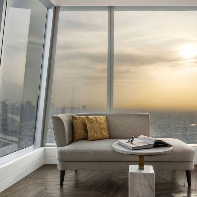 A cozy gray couch with gold pillows sits near a small round table by a large window, overlooking a city skyline at sunset, creating a serene atmosphere.