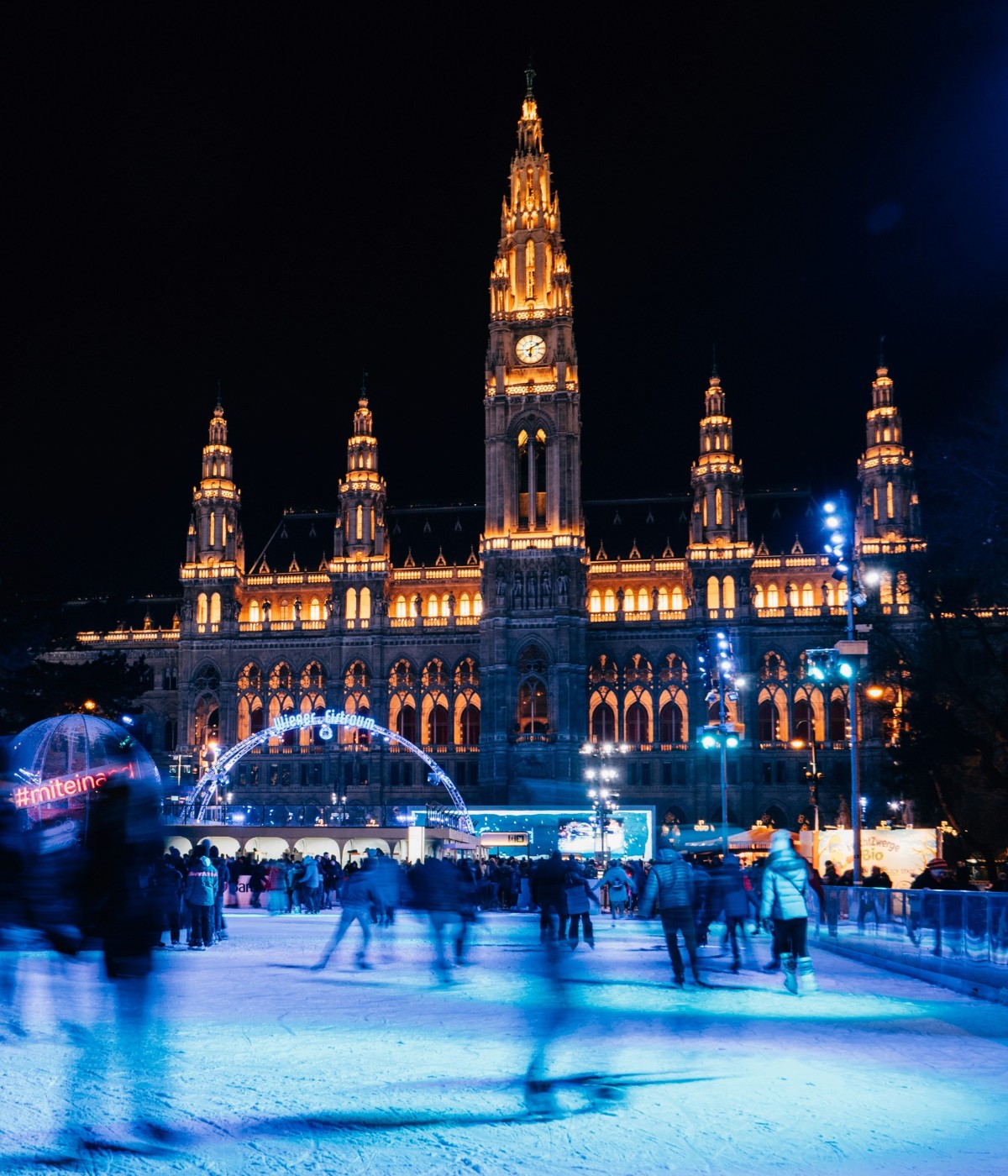 Vienna's illuminated city hall at night, surrounded by an outdoor ice skating rink and bustling visitors.