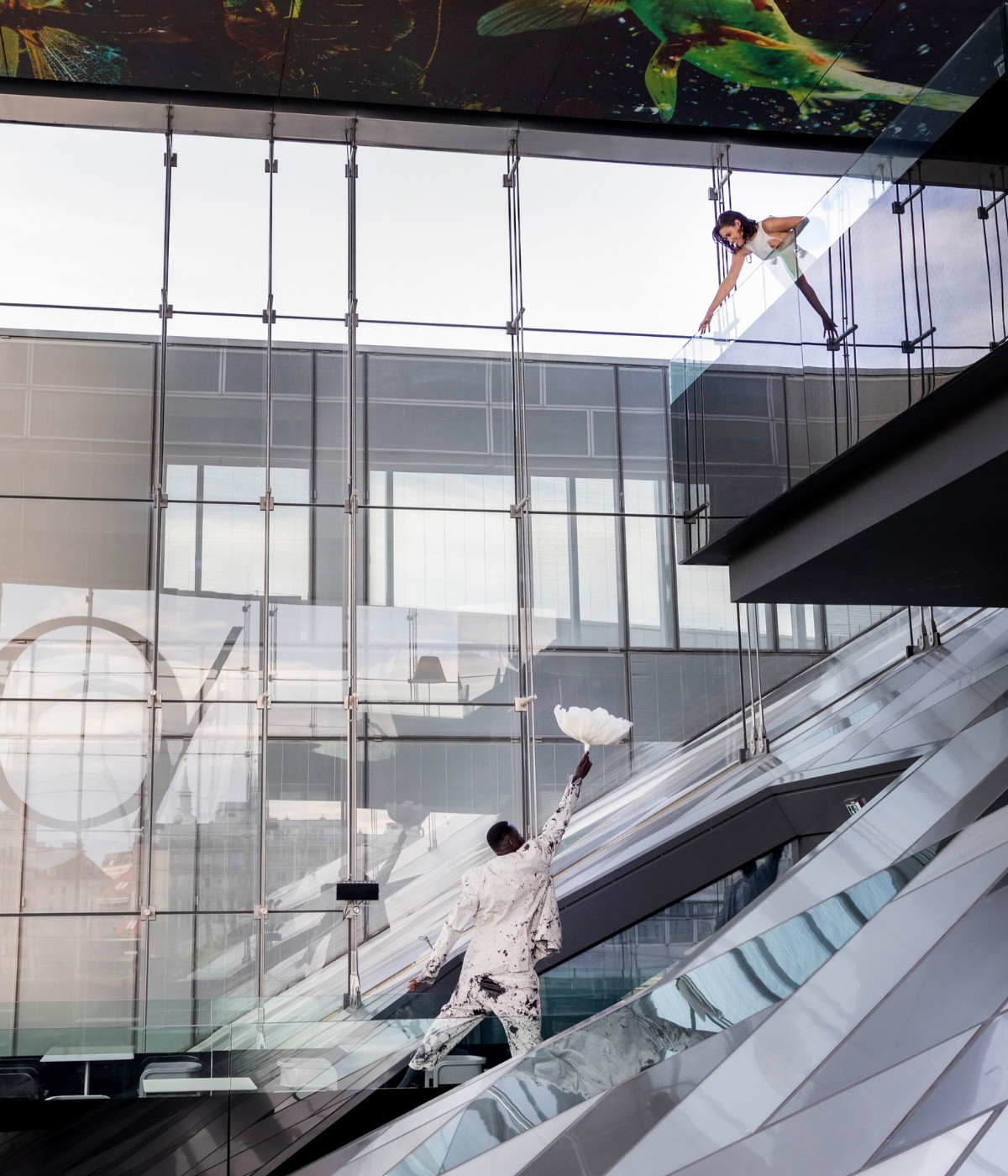A stylish figure in a vibrant outfit poses dramatically on a staircase, surrounded by modern glass architecture and artistic ceiling designs.