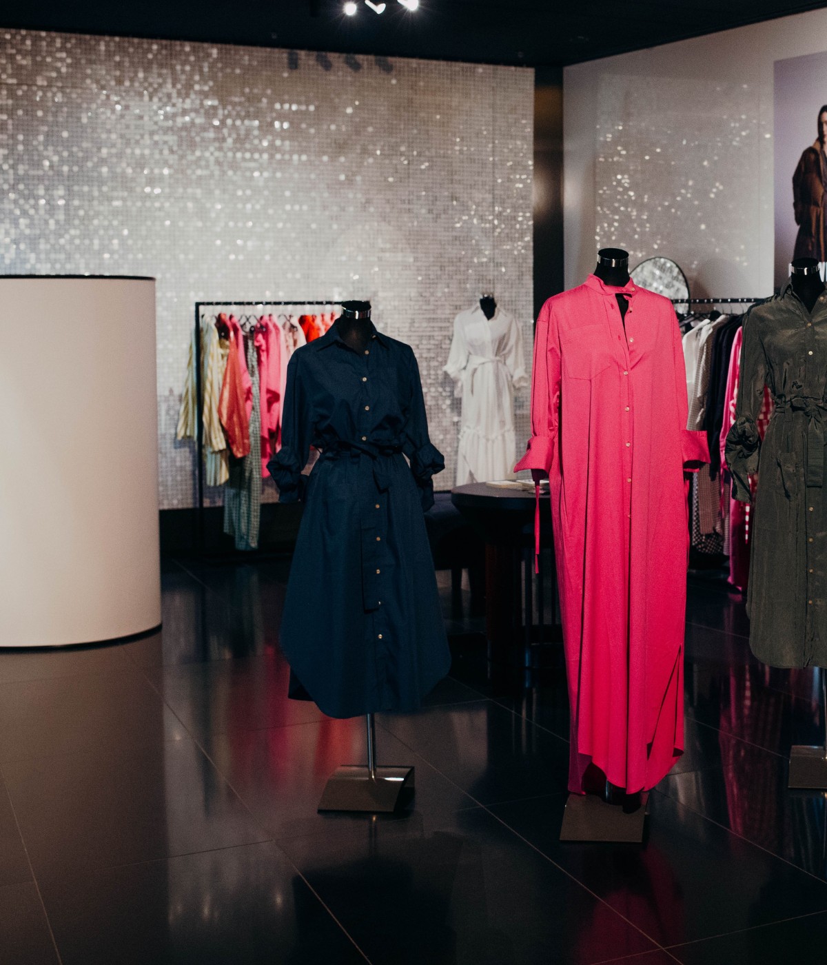A vibrant fashion display featuring a pink dress and a navy shirt dress on mannequins, with colorful clothing hanging in the background.