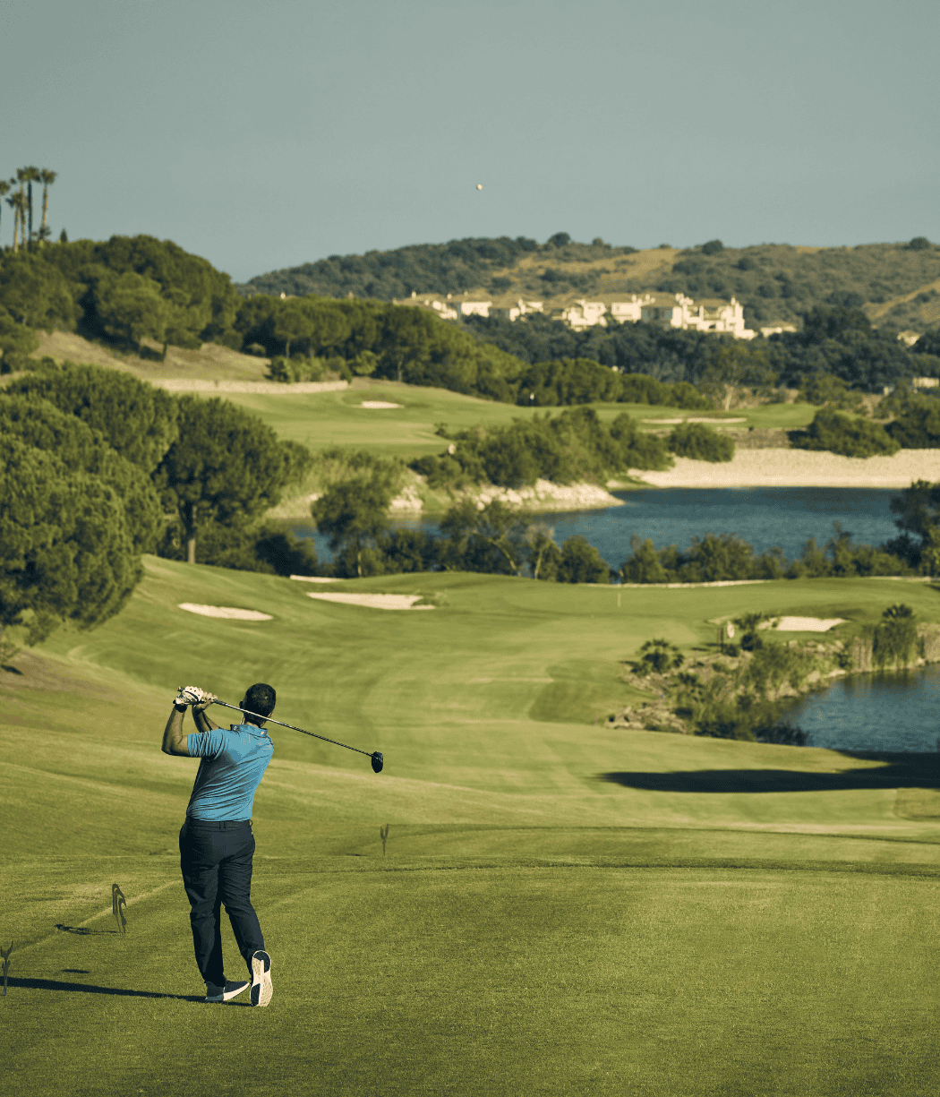 Un golfista con una camiseta azul golpea la bola con un palo en un campo verde y frondoso, con árboles y un lago al fondo bajo un cielo despejado.