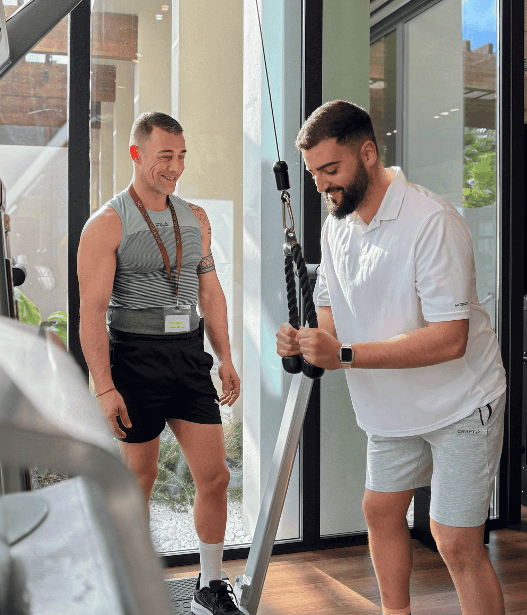 Dos hombres están en un gimnasio; uno muestra cómo se hace un ejercicio con una máquina de poleas mientras el otro observa con atención.