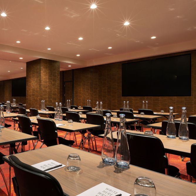 Modern conference room with rows of tables and chairs, set with notepads, glasses, and water bottles. Warm lighting and a large screen create a professional atmosphere.