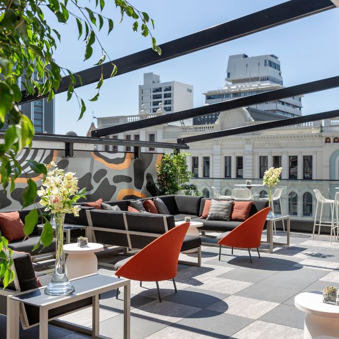 Rooftop lounge with modern seating, orange and gray cushions, and potted plants. Overlooks city buildings, creating a chic and vibrant atmosphere.