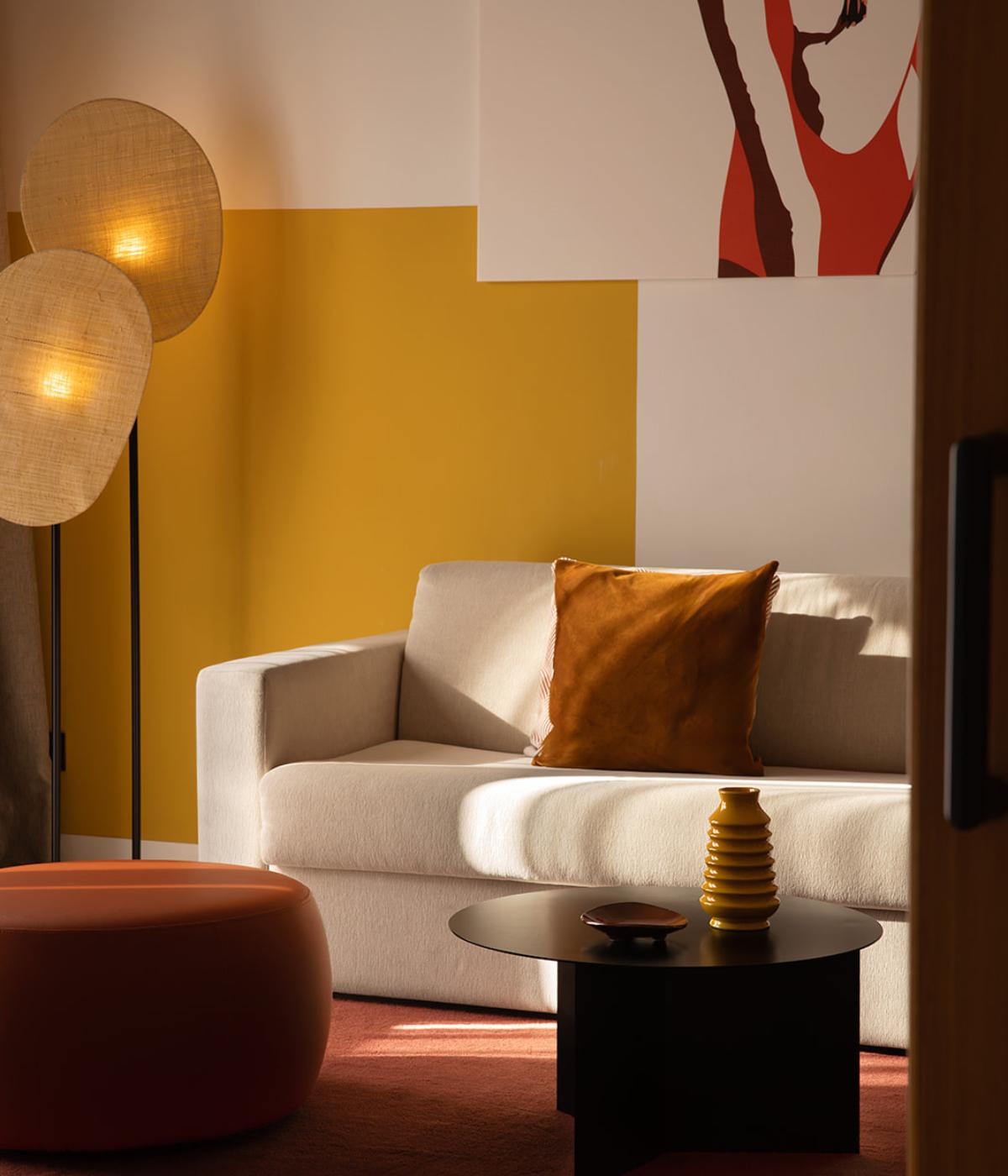 Cozy living room with beige sofa, orange pillow, round black coffee table, modern standing lamps, and mustard-yellow accent wall with artwork.