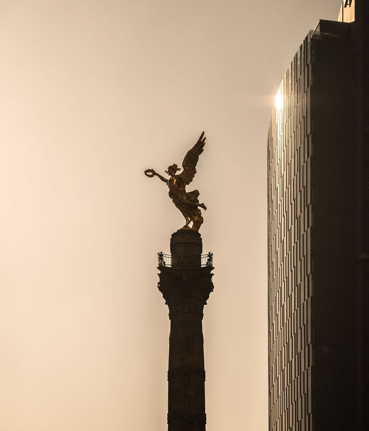 Silhouette of the Angel of Independence monument with modern glass building glowing from sunlight in the background.