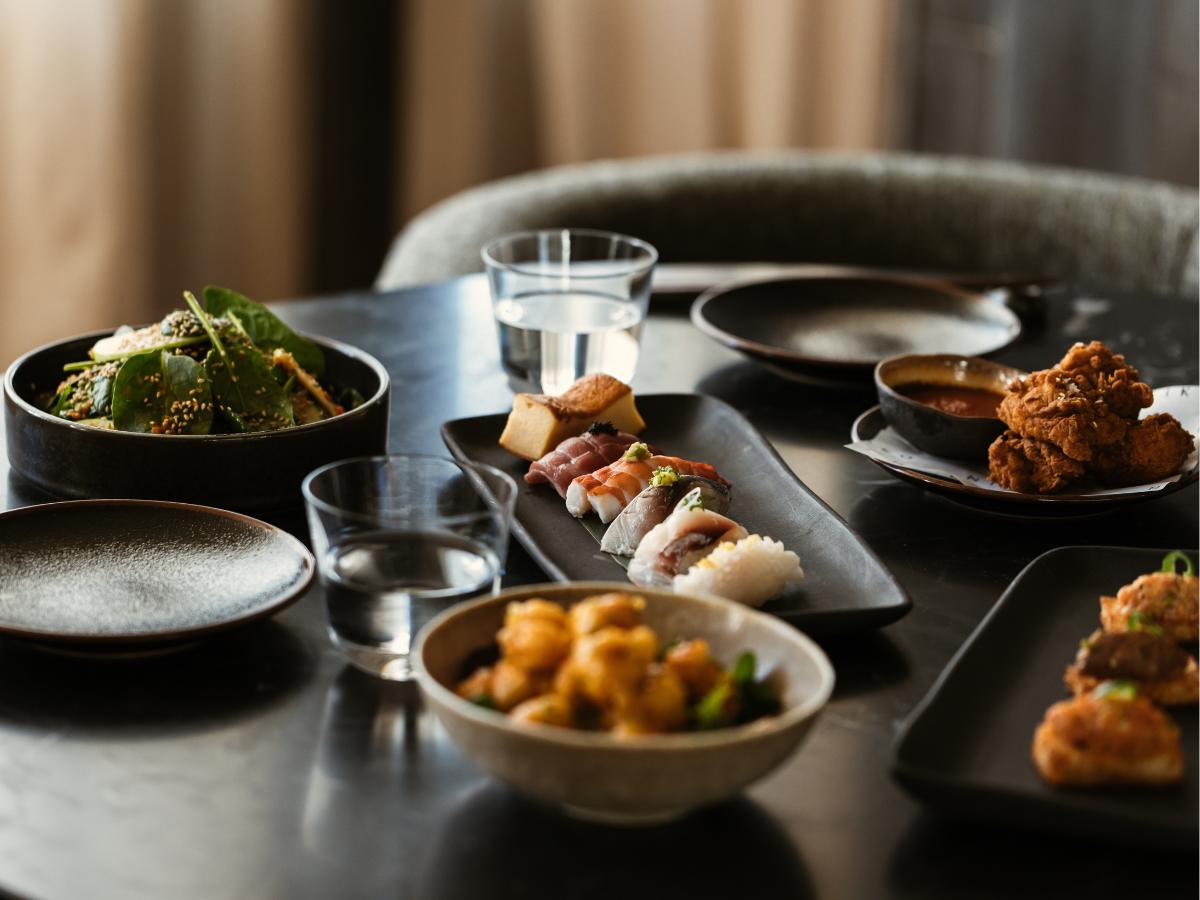Table with assorted dishes including sushi, fried chicken, salad, and two glasses of water in a cozy dining setting.