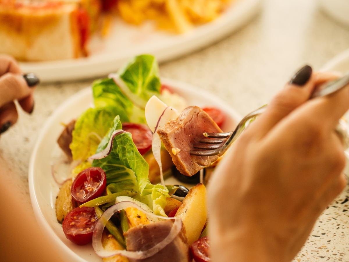 Person holding a fork with seared tuna over a plate of salad including lettuce, cherry tomatoes, onions, and potato wedges.