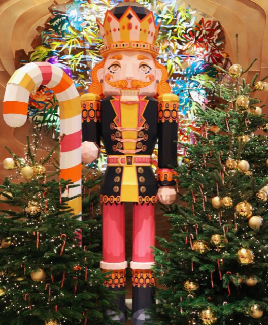 Colorful nutcracker statue with a crown and vibrant attire, holding a candy cane. Flanked by Christmas trees adorned with golden baubles and candy canes.