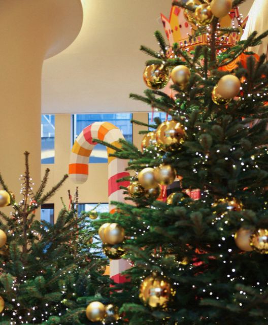 Festive scene with decorated Christmas trees adorned with gold ornaments and lights. Large candy cane in the background adds a playful touch.