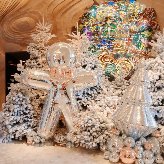 A large silver gingerbread man balloon stands among snow-covered artificial trees with shiny ornaments and a colorful circular decoration in the background.