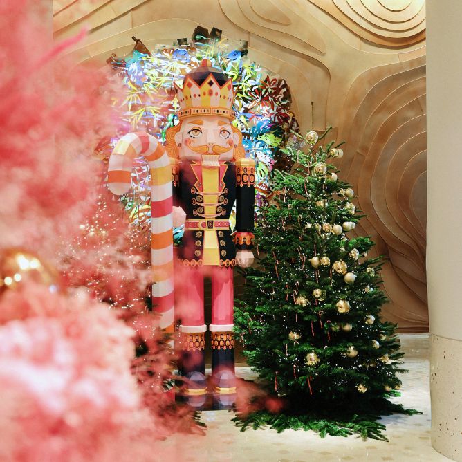 A festive scene features a colorful nutcracker holding a large candy cane, flanked by Christmas trees adorned with golden ornaments, and vibrant decorations.