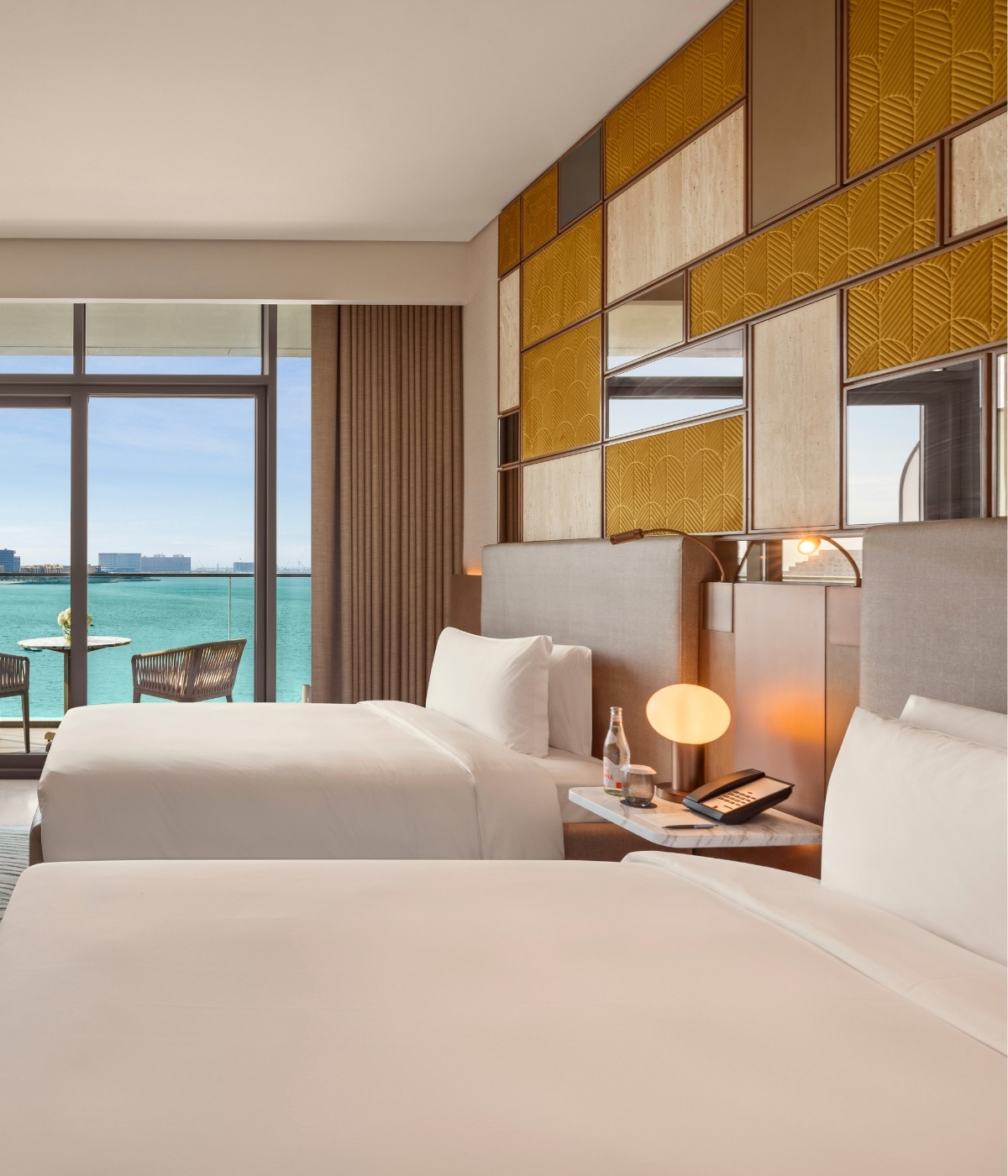 Two neatly made beds are in a modern hotel room. A floor-to-ceiling window offers a stunning ocean view. Warm tones and soft lighting create a serene ambiance.