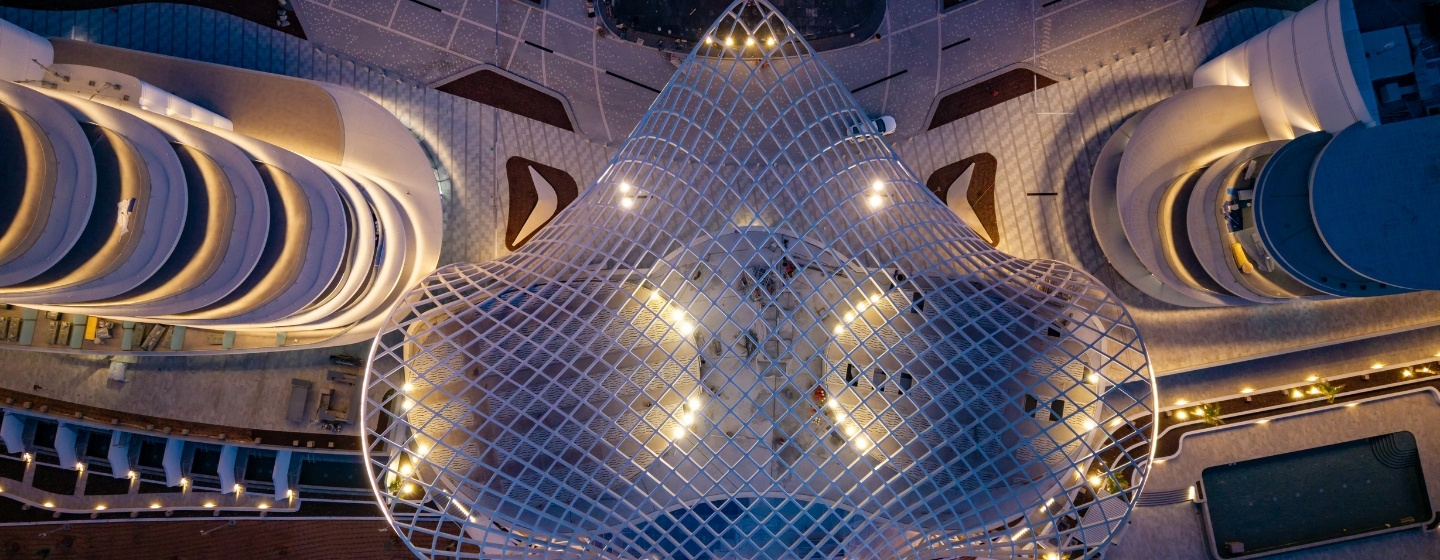 Aerial view of a modern building complex at night, featuring illuminated curved structures and a central net-like roof. The scene conveys a futuristic ambiance.