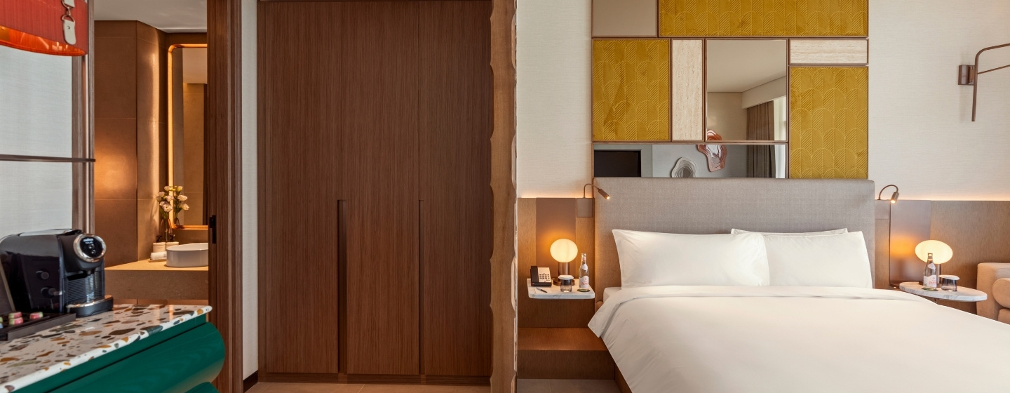 Modern hotel room with a neatly made bed, wooden closet, and mirrored wall accents. Warm lighting from bedside lamps creates a cozy ambiance. A coffee machine sits on a green counter, adding elegance.