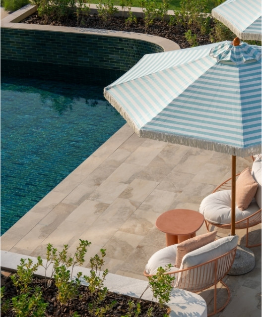 http://Poolside%20scene%20with%20turquoise%20water,%20beige%20tiles,%20two%20cushioned%20chairs,%20a%20round%20table,%20and%20a%20large%20striped%20umbrella.%20Greenery%20adds%20a%20serene%20touch.