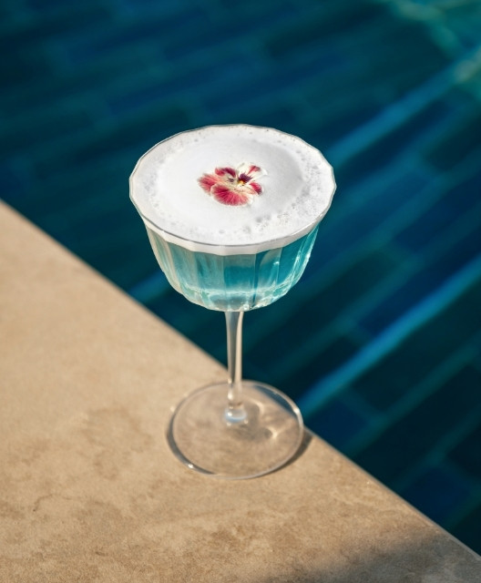 http://A%20frothy,%20light%20blue%20cocktail%20in%20a%20clear%20glass%20with%20a%20purple%20flower%20garnish,%20set%20against%20a%20pool's%20serene%20blue%20water,%20conveys%20a%20refreshing%20vibe.