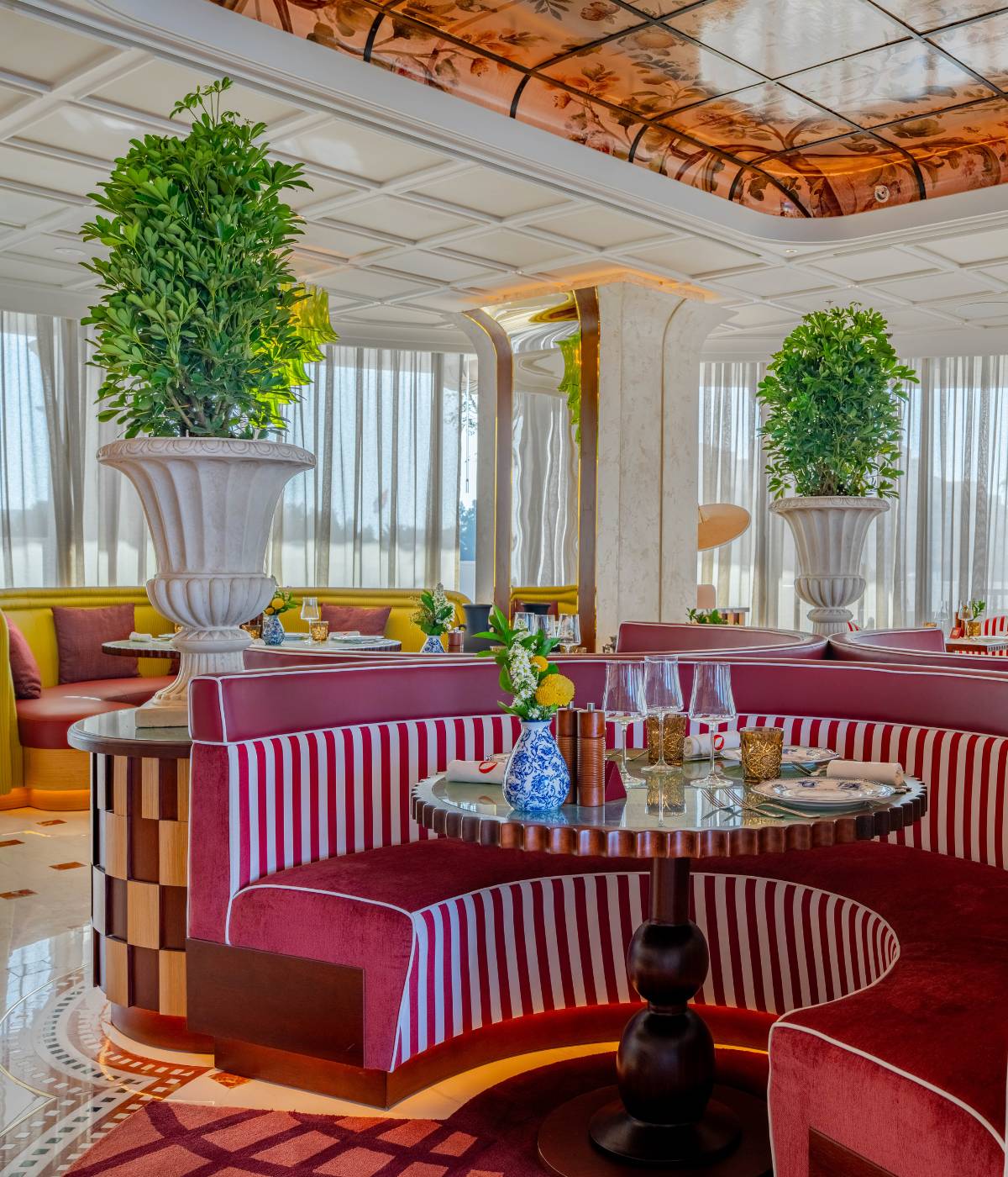 Luxurious restaurant interior with burgundy and white striped booths, large potted plants, elegant table settings, and natural light filtering through sheer curtains.