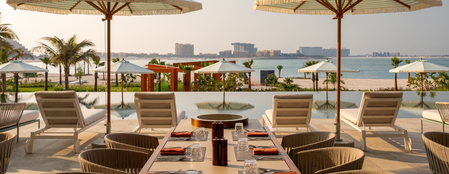 Chic outdoor dining area overlooking a serene beach, with sun loungers and umbrellas, surrounded by palm trees and a tranquil water feature.