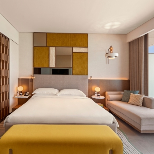 http://A%20modern%20hotel%20room%20with%20a%20double%20bed,%20geometric%20headboard,%20mustard%20and%20beige%20accents,%20cozy%20lighting,%20and%20a%20plush%20sofa,%20creating%20a%20warm,%20inviting%20ambiance.