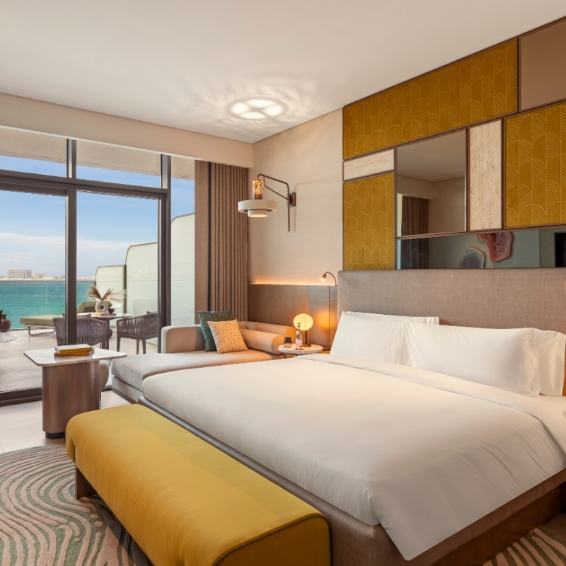 Bright, modern hotel room with a king bed, a seating area, and ocean views through large glass doors. Elegant decor in warm tones.