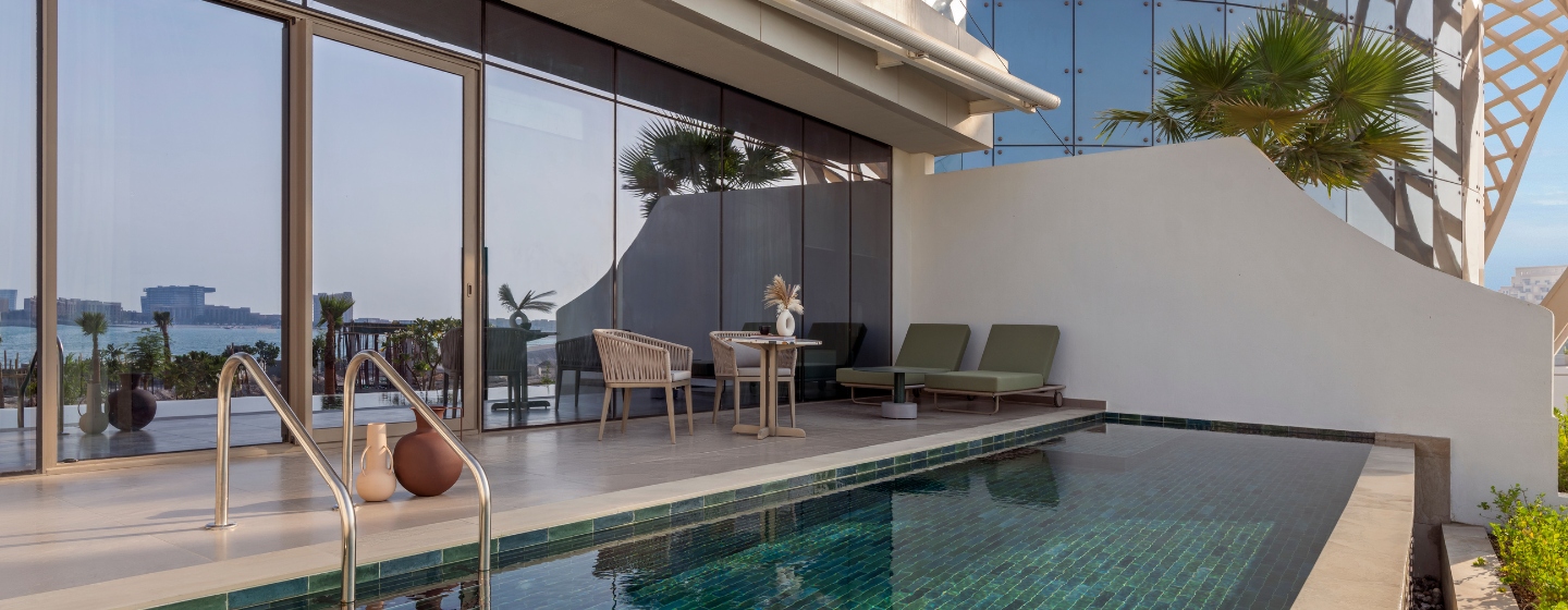 Luxurious poolside patio with modern furniture, including two lounge chairs and a small table. Glass doors open to a serene ocean view, creating a tranquil atmosphere.