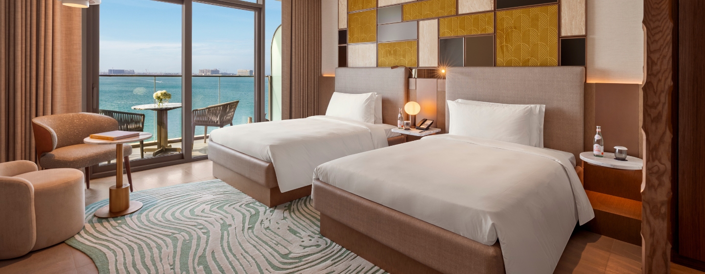 Modern hotel room with two beds featuring white linens, an abstract rug, and a patterned accent wall. A balcony offers a serene ocean view.
