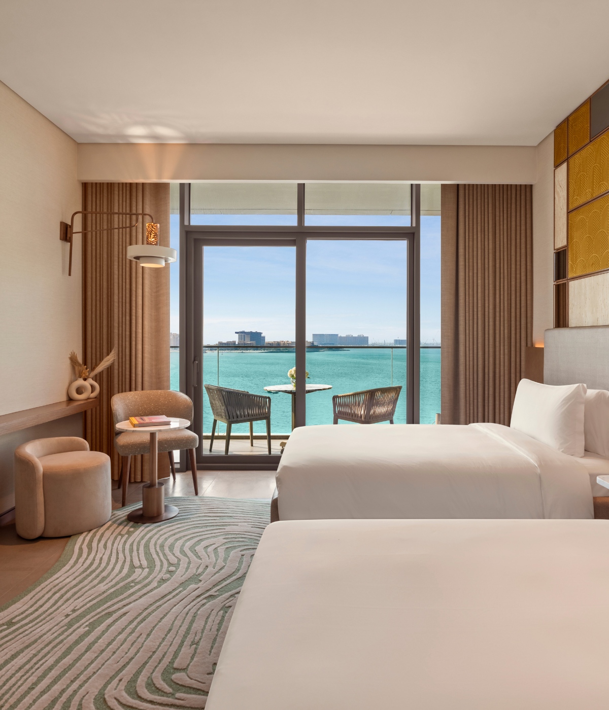 Hotel room with two beds, modern decor, and a balcony overlooking a tranquil sea. Sunlit room, beige tones, and two chairs on the balcony create a serene atmosphere.