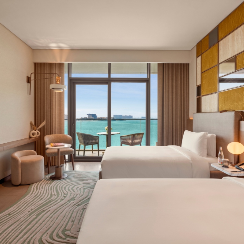 Modern hotel room with two white beds, a patterned rug, and a wall of gold accents. A balcony reveals a calm turquoise ocean under a clear blue sky.