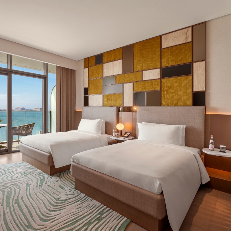 Modern hotel room with two neatly made twin beds. A geometric patterned wall adds warmth, large window shows a sea view, creating a serene atmosphere.