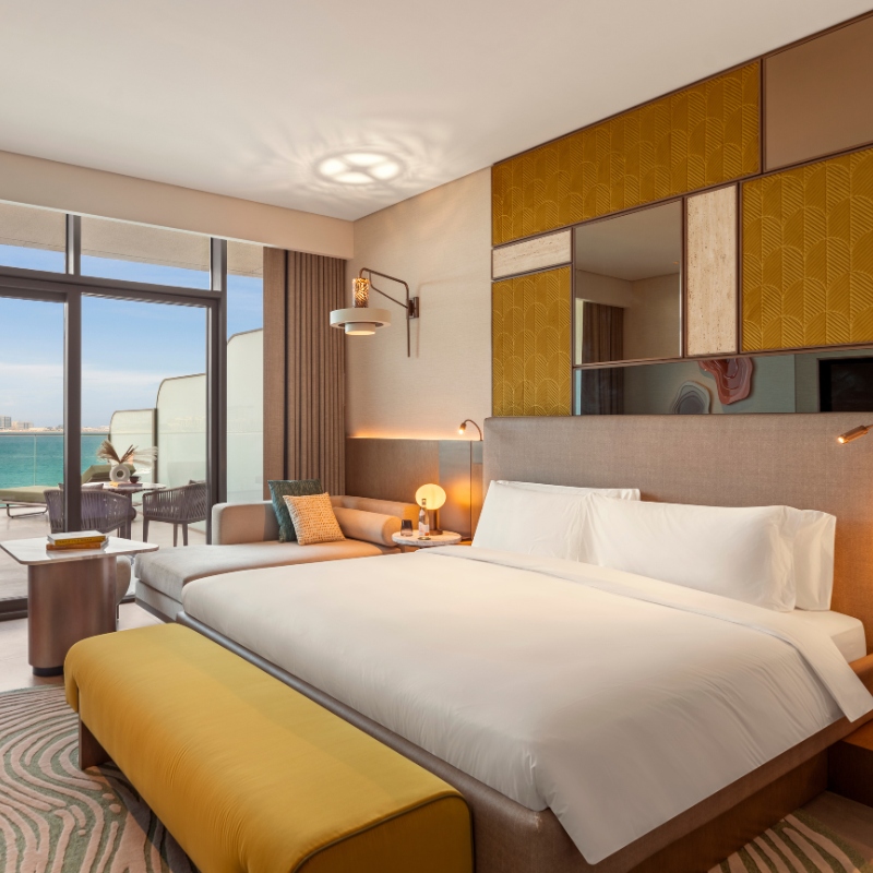 Modern hotel room featuring a large bed with white linens, a yellow bench, and ocean-view balcony. Cozy, elegant design with warm lighting.
