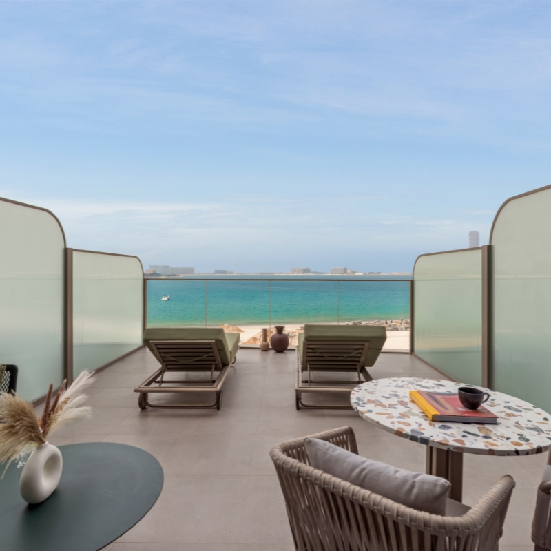 Serene balcony view with two lounge chairs facing a blue ocean under a clear sky. A cozy seating area with a small table and decor in the foreground.