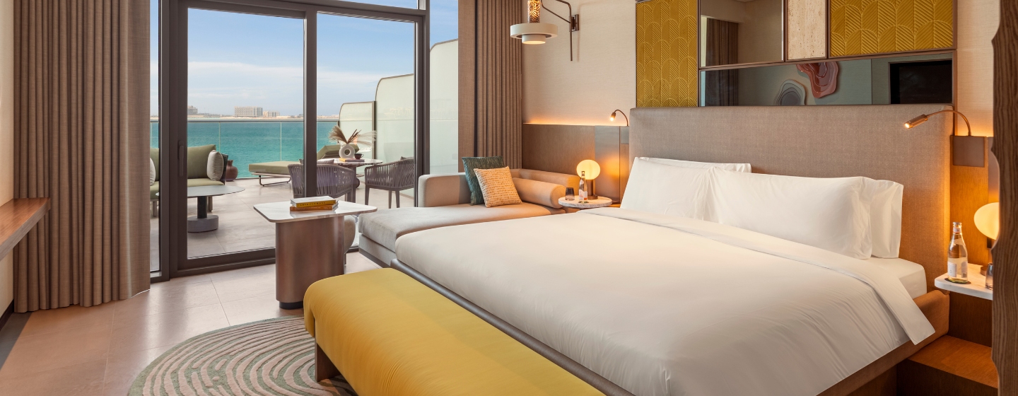 Modern hotel room featuring a large bed with white linens, a yellow bench, and ocean-view balcony. Cozy, elegant design with warm lighting.