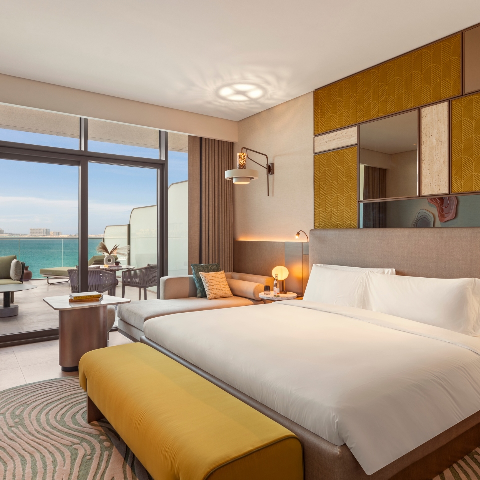 Modern hotel room featuring a large bed with white linens, a yellow bench, and ocean-view balcony. Cozy, elegant design with warm lighting.